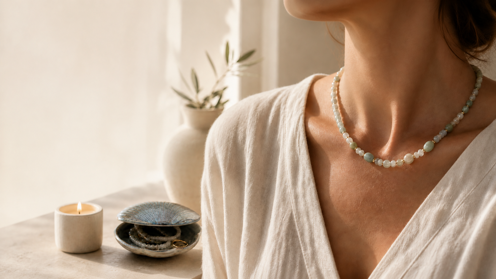 Woman wearing a pale gemstone necklace beside a candle and shell jewellery box in warm window light